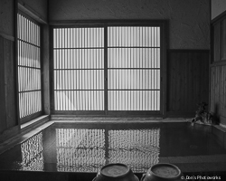 Black and white photograph of an indoor hot spring with light shining through the closed shoji door reflecting off the still water.